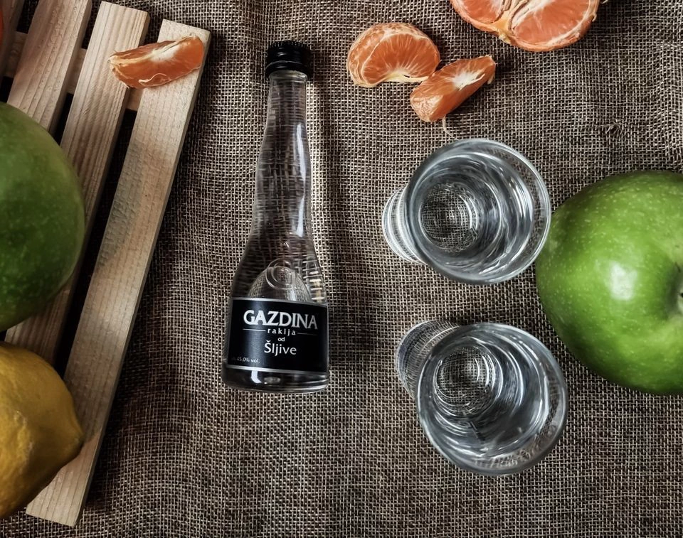 Boca rakije i dvije čaše stoje na jutenoj tkanini, okružene voćem (zeleni jabuka, limun, mandarina). Tekst: "GAZDINA rakija od Šljive".