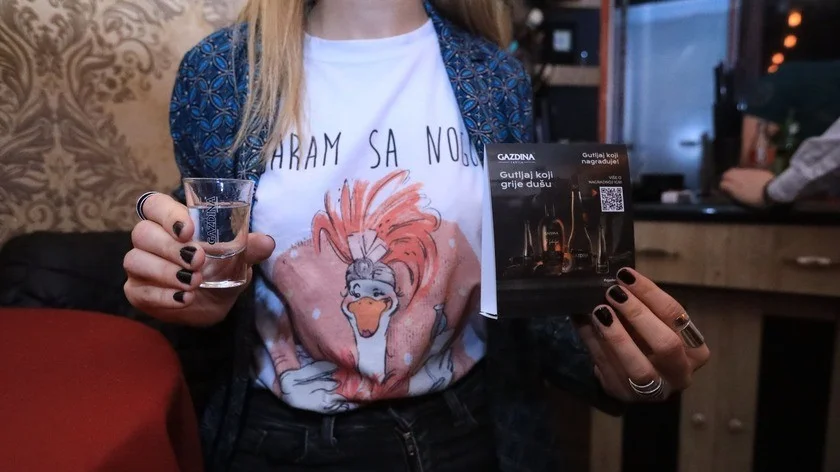 Osoba drži čašu vode i prospekt u kafiću. Majica sadrži tekst "HARAM SA NOGU".