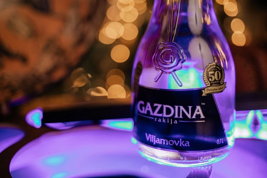 Boca rakije GAZDINA svijetli u ljubičastom osvjetljenju; u pozadini su mutna svjetla. Tekst: "GAZDINA rakija Viljamovka 0.7L" i "Prirodna i zrela 50 godina".